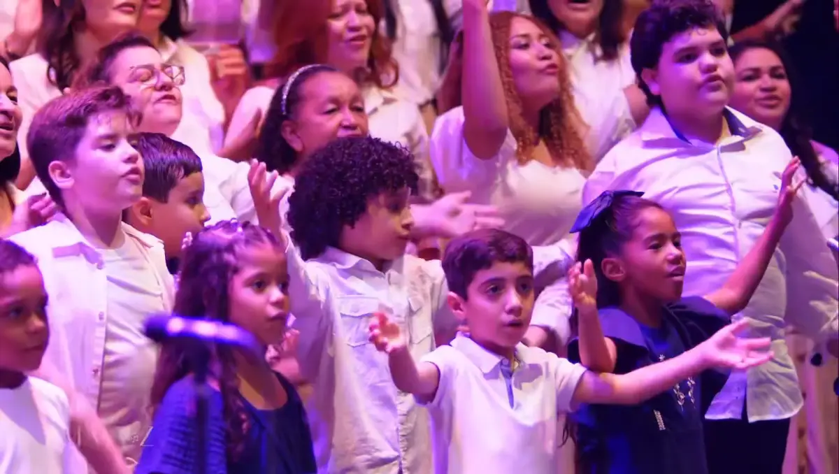 Crianças do coral cantando, de camisa branca, com adultos ao fundo